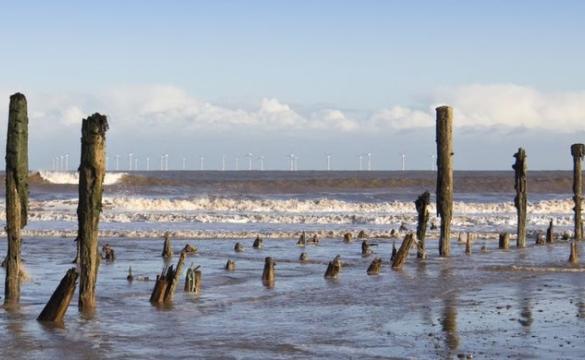 Remnants of a pier sticking out of the sea shore with wind turbines in the distance