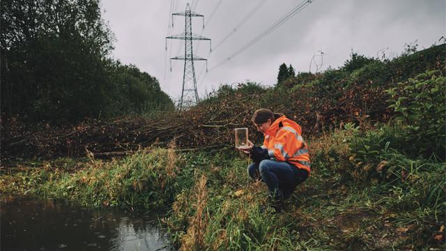 Person wearing orange PPE jacket taking a water sample from a pond with an electricity pylon in the background