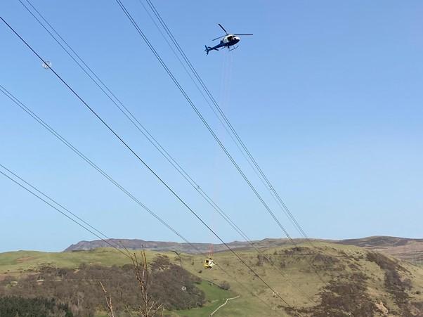 NGHelicopterLiveLineWork.jpg Helicopter hovering over electricity overhead lines with basked carrying engineers suspended below