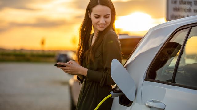 PersonChargingEV_640x360.jpg Person holding a mobile device leaning against electric vehicle while it's charging